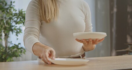 A woman in a white sweater is arranging white ceramic plates on a wooden table in a bright kitchen.