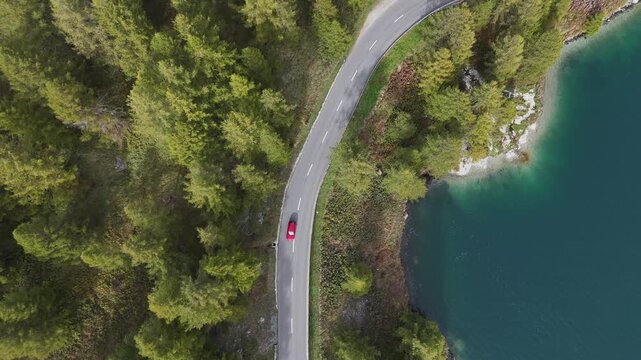 Aerial view of a vibrant red car traverses a winding road alongside a serene lake, contrasting with the lush green forest, Sils im Engadin Segl, Grisons, Switzerland.