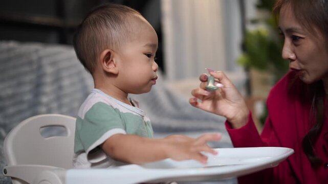 Mother feeding her cute asian toddler boy in high chair
