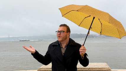 A Caucasian man with a quirky yellow umbrella contemplates seaside showers, evoking Umbrella Day and World Maritime Day