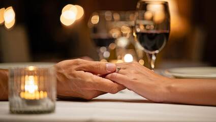 Close-up of two people holding hands across a candlelit dinner table with soft bokeh lights and glasses of red wine, creating a warm romantic mood