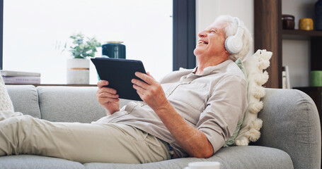 Obraz premium Laughing, music and tablet with old man on sofa in living room of home for subscription streaming. App, funny and headphones with happy senior person in apartment for audio or sound entertainment