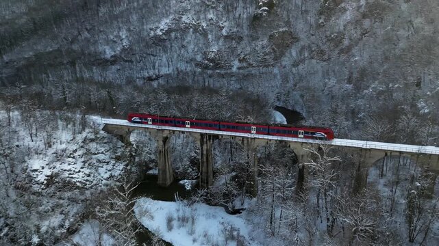 Valjevo, Serbia - 27 October 2025: Aerial view of a vibrant red passenger train traversing a snow-laden bridge amidst a forest of trees covered in white snow.