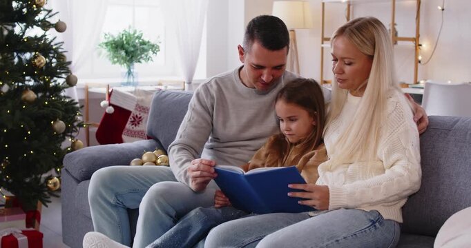 Family reading a Christmas book at home. Parents sit beside daughter on the sofa by the tree, warm lights and gifts around them as the child follows the pages. Warm family holiday at home.