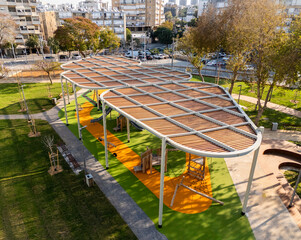 Aerial view of a modern playground with vibrant green synthetic turf and winding orange pathways, under the shade of an innovative wooden and metal canopy, Bat Yam, Center District, Israel.