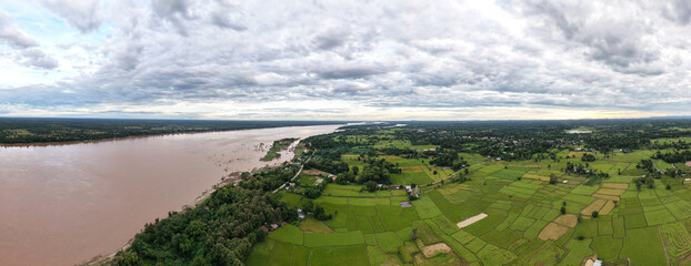 Panoramic Aerial View of Mekong River and Rice Fields flowing beside vibrant green rice paddies and rural villages under dramatic cloudy sky. 