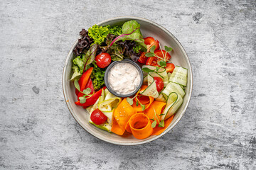 Healthy fresh vegetable salad with cucumber ribbons, carrot curls, lettuce, cherry tomatoes, bell pepper, microgreens, and creamy dipping sauce served in a modern bowl