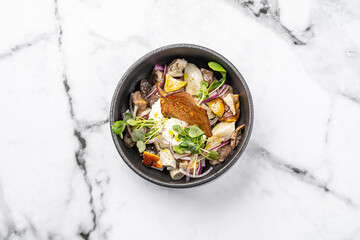 Gourmet salad with assorted mushrooms, creamy cheese, red onion, microgreens, and crispy bread served in a black bowl on marble background
