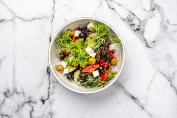 Fresh Greek salad with feta cheese, olives, lettuce, cucumber, tomato, and herbs served in a ceramic bowl on marble background