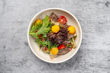 Fresh gourmet salad with slices of beef, cherry tomatoes, crispy lettuce, cucumber, fried potato balls, and mixed greens served in a ceramic bowl on gray rustic background
