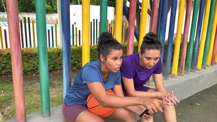 Two young women in sportswear sitting outdoors using a smartphone and discussing strategies - Powered by Adobe