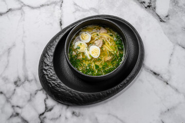 Traditional chicken noodle soup with boiled egg slices, fresh dill, and vegetables served in a black ceramic bowl on marble table