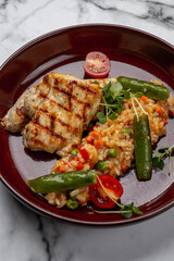 Grilled chicken breast served with vegetable risotto, green peas, cherry tomatoes, and microgreens on a red ceramic plate on marble background