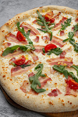 Freshly baked pizza with crispy bacon, arugula leaves, melted mozzarella cheese, and cherry tomato slices on wooden board on gray stone background