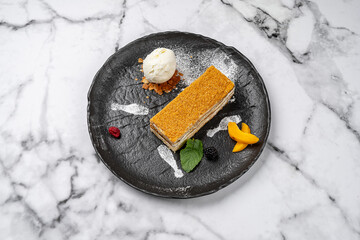 Traditional layered honey cake Medovik served with vanilla ice cream scoop, fresh fruits, berries and mint on black plate with marble background