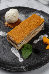 Traditional layered honey cake Medovik served with vanilla ice cream scoop, fresh fruits, berries and mint on black plate with marble background