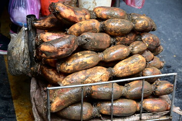 Stacked lotus roots with brown soil-stained skin on metal wire rack, plastic bags visible in blurred market background, fresh produce display, Taiwan traditional market scene.