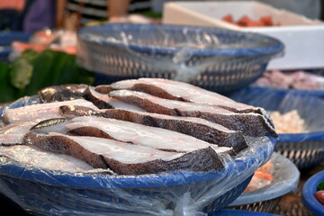 Blue plastic basket filled with stacked white fish fillet slices showing dark skin edges, covered with clear plastic wrap, silver basket with items visible behind, market vendor display, Taiwan.