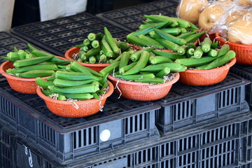 Five red plastic baskets filled with fresh green okra (lady's finger) on black plastic crates, light-colored round fruits visible in basket behind, vendor display at market, Taiwan.