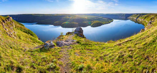 A breathtaking panoramic view of the Dniester River canyon near Subich, Ukraine. Mossy rocks are in the foreground, overlooking the calm blue water and green forested hills under a bright sun.