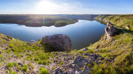 A breathtaking panoramic view of the Dniester River canyon near Subich, Ukraine. Mossy rocks are in the foreground, overlooking the calm blue water and green forested hills under a bright sun.
