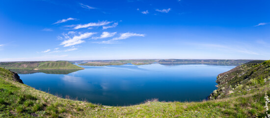 A beautiful aerial panoramic landscape of the Bakota Bay on the Dniester River in Ukraine. The wide, calm blue water is surrounded by large green hills under a bright spring sky.