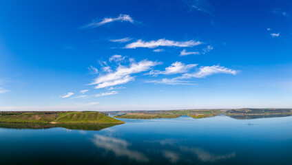A beautiful aerial panoramic landscape of the Bakota Bay on the Dniester River in Ukraine. The wide, calm blue water is surrounded by large green hills under a bright spring sky.