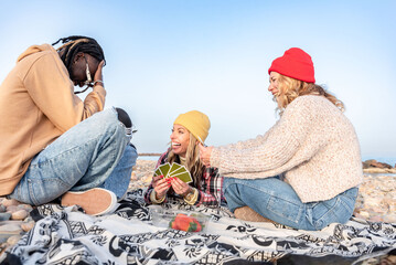 Group of diverse happy friends playing cards and enjoying a picnic on a rocky beach