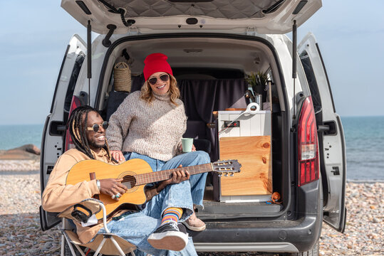 Multiracial couple playing guitar and having fun at beach with camper van