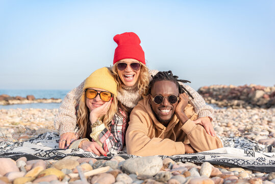 Happy multiracial diverse friends in sunglasses lying on a pebble beach smiling at camera