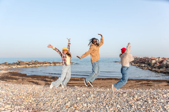 Three diverse friends having fun jumping on a pebble beach by the sea