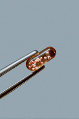 Translucent amber capsule held by tweezers on neutral background, macro pharmaceutical supplement and clinical research detail
