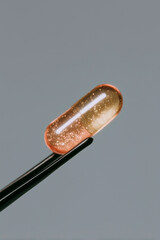 Translucent amber capsule held by tweezers on neutral background, macro pharmaceutical supplement and clinical research detail