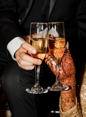 Two glasses with sparkling wine in hands of newlyweds in the wedding day. The bride and groom clink glasses with champagne. Close-up of hands with henna patterns holding festive glasses of champagne