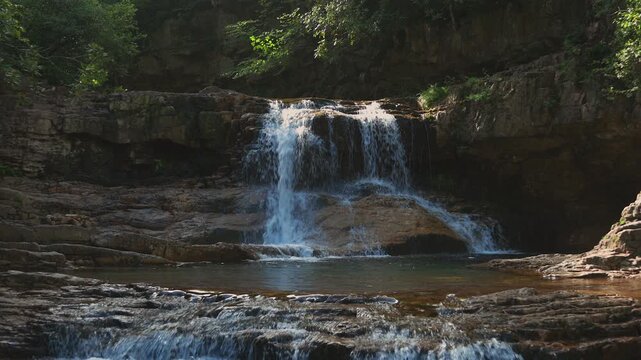 Sunlight hits St. Mary's Falls as it cascades over a rocky ledge into a tranquil pool. Filmed on a summer morning in St. Mary's Wilderness, Virginia (George Washington NF).