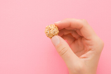 Young adult woman hand fingers holding white chocolate truffle with nut sprinkles on light pink background. Pastel color. Sweet snack. Closeup. Front view.