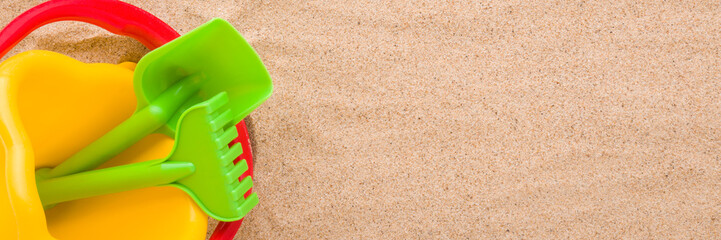 Colorful bucket with spade and rake on light brown sea sand background. Closeup. Playing plastic toys for baby. Wide banner. Flat lay. Empty place for text. Top down view.