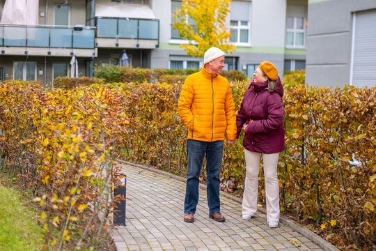 Happy senior couple enjoying walk together on paved autumn path, modern apartment buildings, man in yellow jacket, woman in burgundy coat, active retirement, and beauty aging gracefully side by side