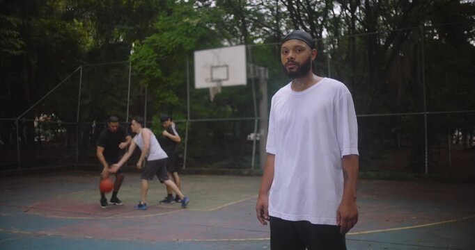 African descent man wearing durag standing on basketball court observing teammates play, showing calmness, confidence, and connection to the team during a casual urban game