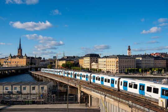 Elevated view of blue metro train crossing railway tracks in the urban area of Stockholm city near Gamla Stan, showing modern transport infrastructure and skyline under soft daylight atmosphere