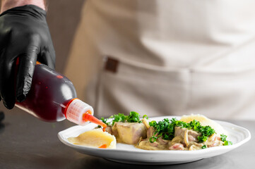 A chef in a professional kitchen is plating a gourmet dish, drizzling a red sauce from a squeeze...