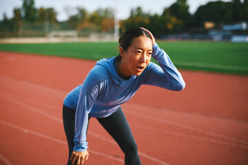 Exhausted young Asian woman stands hunched over with her hand on her head, catching her breath after a strenuous workout on a sunny outdoor running track.