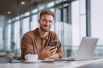 Young male professional using a mobile phone, smiling at camera while working on a laptop in a contemporary office setting