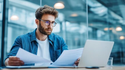 Obraz premium Young businessman in glasses reading papers while working with laptop on a desk in a contemporary office setting