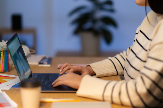 Crop unrecognizable woman in casual sweater sitting at table with laptop and working online on project from home during evening time