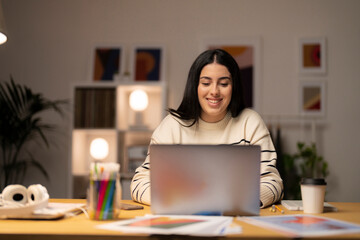 Young female freelancer in striped sweater sitting at table with laptop working on startup strategy in creative workspace looking at camera