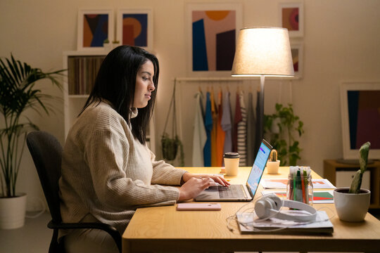 Positive young Arab woman in casual sweater sitting at table with laptop and working online on project from home during evening time