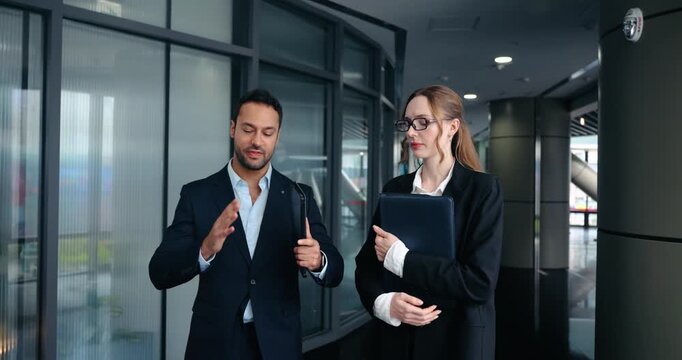 Confident experienced boss explains project details and gives constructive feedback to focused female employee while walking together in modern office corridor