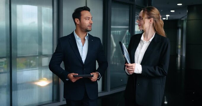 Two positive business colleagues man and woman walk through modern office hallway smiling and having professional conversation