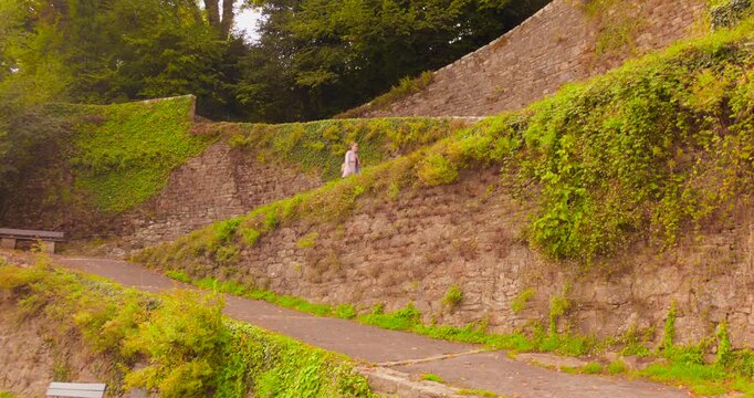 A woman climbs the stone-walled rampes du Loch, from the historic Saint-Goustan port up to the town of Auray. Features ancient stone, ivy, and lush vegetation.